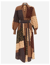 A long-sleeved patchwork midi dress with a waist tie belt, featuring various geometric and polka dot patterns in shades of brown and beige, shown against a light grey background.