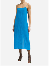 A woman modeling a bright blue midi-length slip dress with thin straps and a gathered, flowing silhouette, paired with brown strappy high-heeled sandals against a light gray studio background.