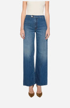 A model wearing a pair of wide-leg blue denim jeans with a faded wash, paired with a cream top and brown pointed-toe heels, set against a light grey background.