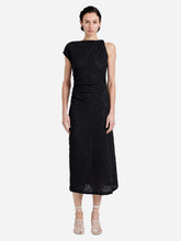 A model wearing a black sleeveless midi dress with a textured lace-like pattern, draped neckline, and gathered waist, paired with beige strappy high-heeled sandals against a light gray studio background.
