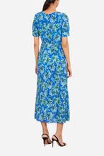 A woman viewed from the back wearing a short-sleeve midi dress with a blue, green, and white floral print, paired with black high-heeled sandals, against a plain white background.