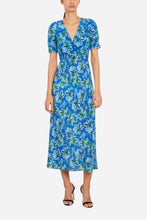 A woman modeling a short-sleeve midi dress with a blue, green, and white floral print, paired with black high-heeled sandals, against a plain white background.