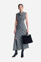 A model stands against a light gray studio background wearing a sleeveless gray plaid midi dress with a high draped neckline, a side slit, and black pointed ankle boots. She is holding a black tote bag.