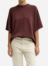 A loose short-sleeve crewneck top in a muted burgundy-brown color, paired with light beige pants, against a plain light gray studio background.